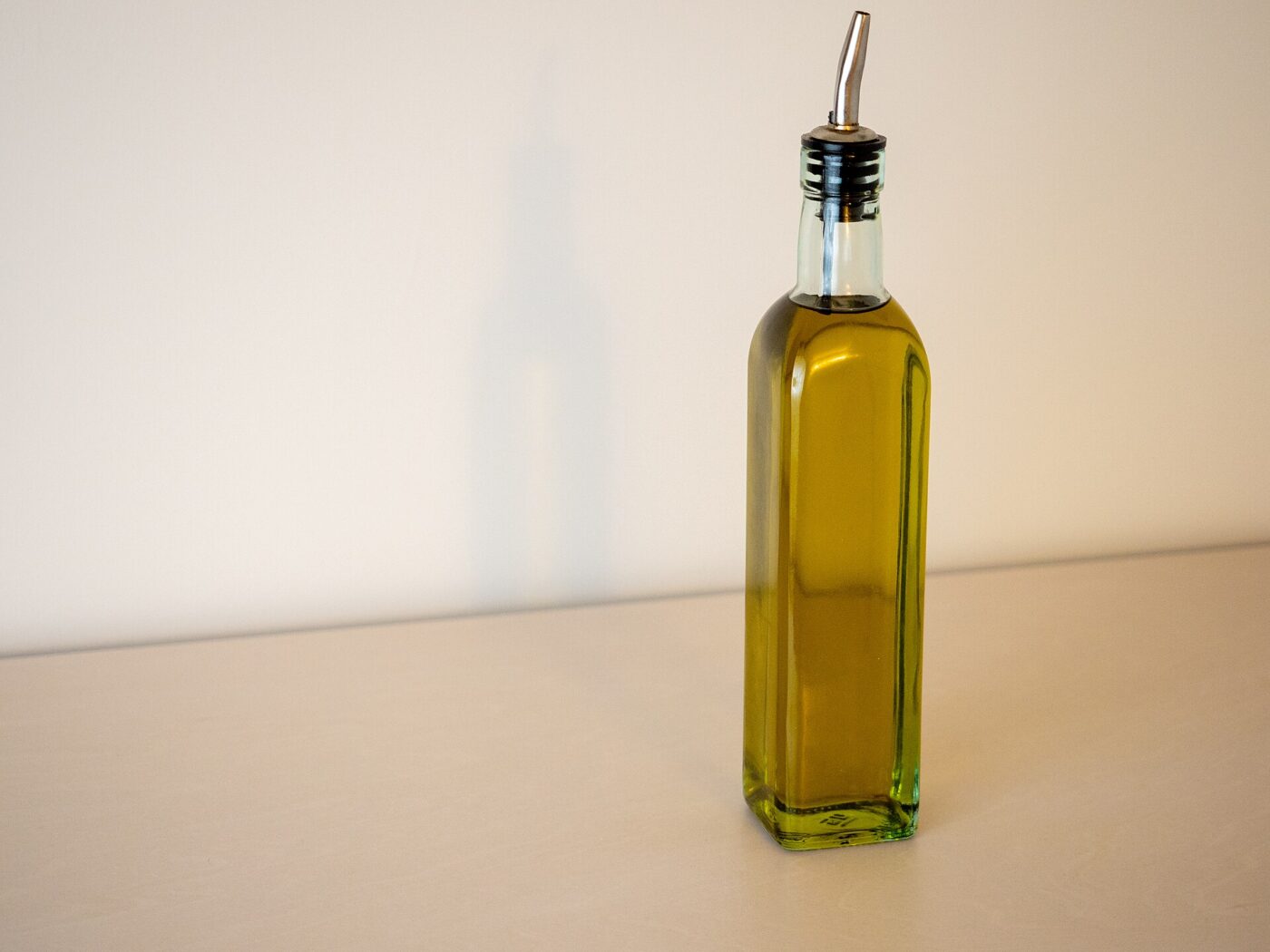 A square glass bottle of green-gold olive oil fitted with a stainless steel pour spout