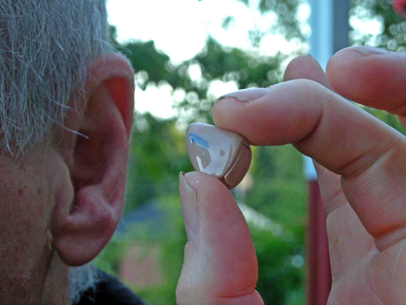 An in-the-ear hearing aid held between fingertips next to an older man's ear