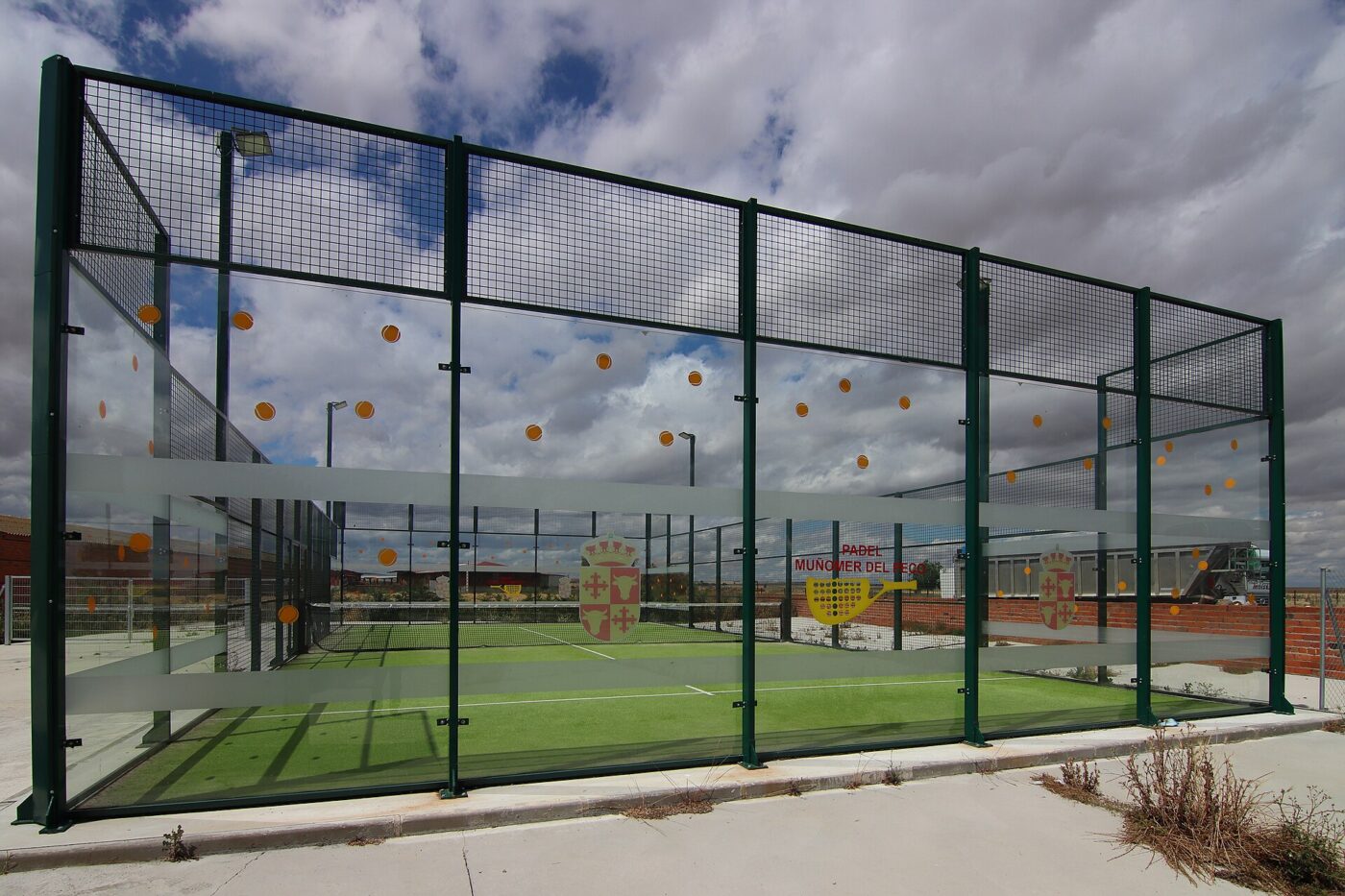 An outdoor padel court enclosed by glass back walls and wire mesh fencing, with green artificial turf