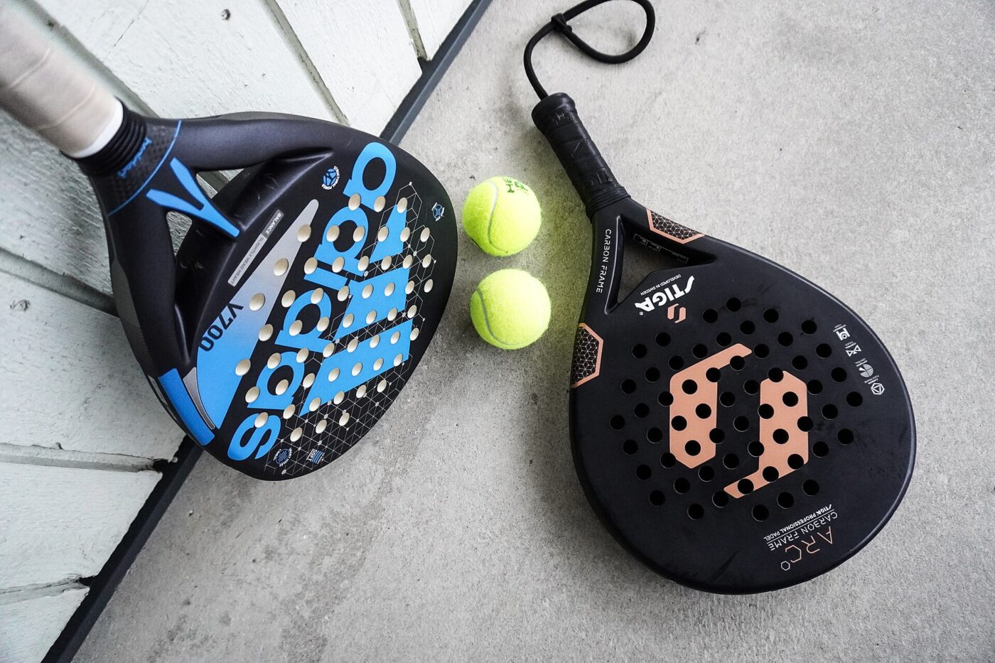 Two padel rackets with perforated hitting faces lying beside two yellow balls on a concrete floor