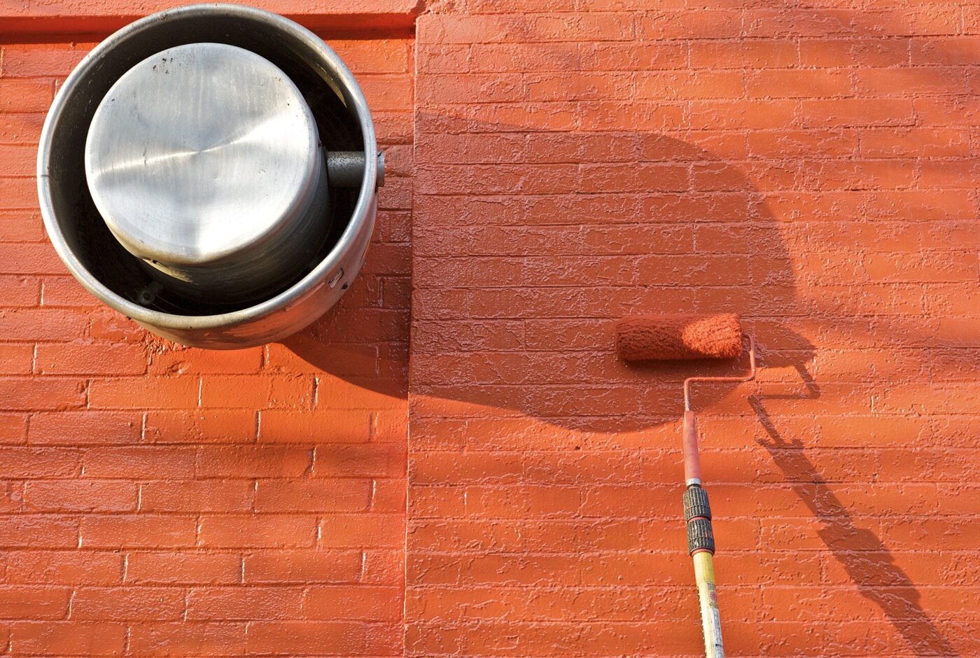 Paint roller on an extension pole coating a brick wall in orange paint beside a vent