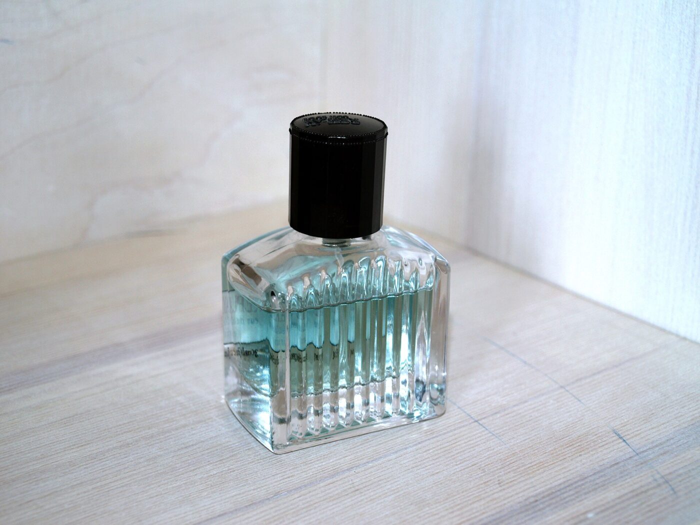 Small ribbed glass perfume bottle with pale blue liquid and a black cap on a wooden surface