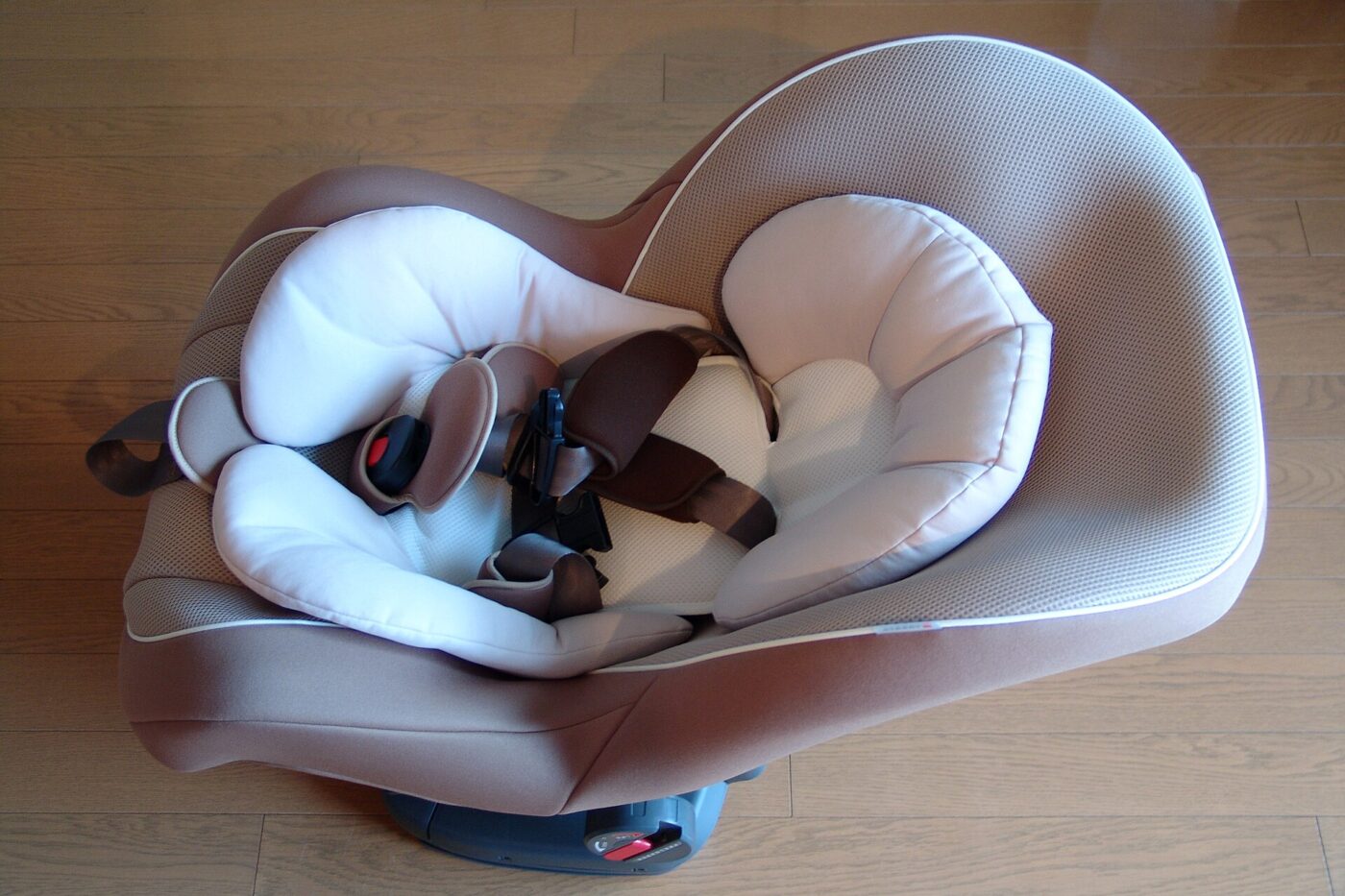 An empty padded child car seat with harness straps, seen from above on a wood floor.