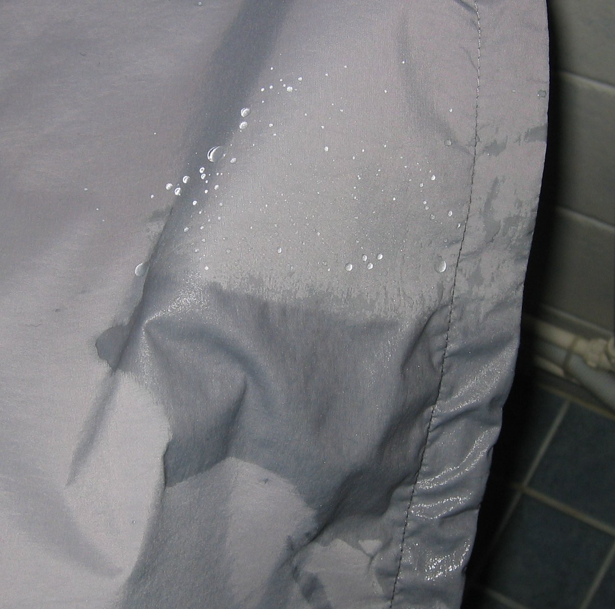 Water droplets bead on a shell jacket's fabric beside a darker patch where the surface has wetted out