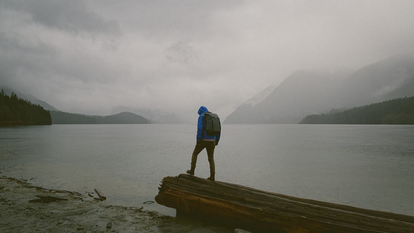 A hiker in a blue hooded rain jacket stands on a log at the edge of a misty lake