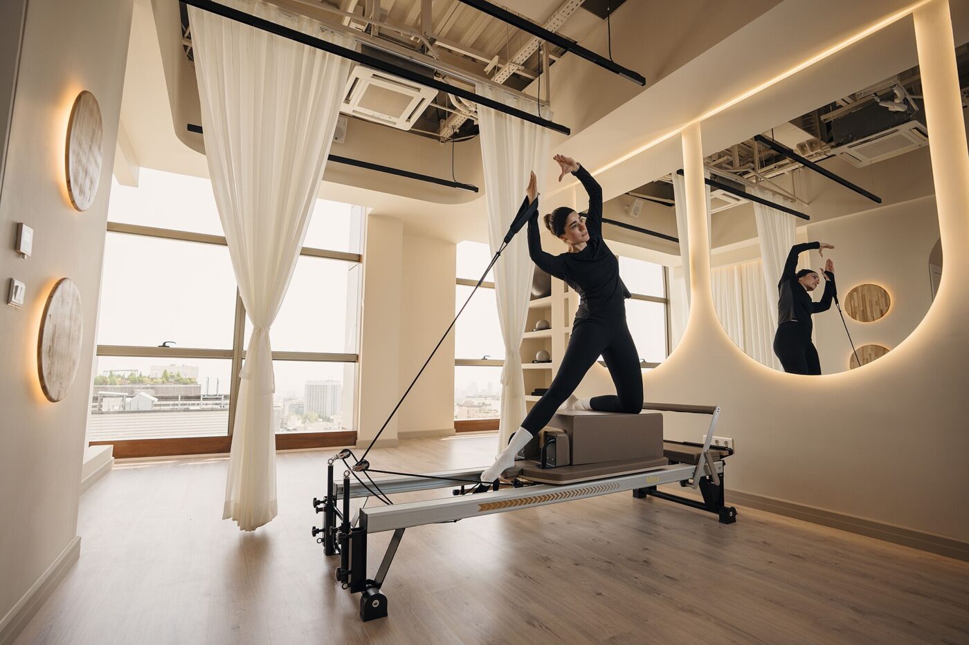 A woman kneels on a Pilates reformer and pulls a strap in a bright studio with white curtains