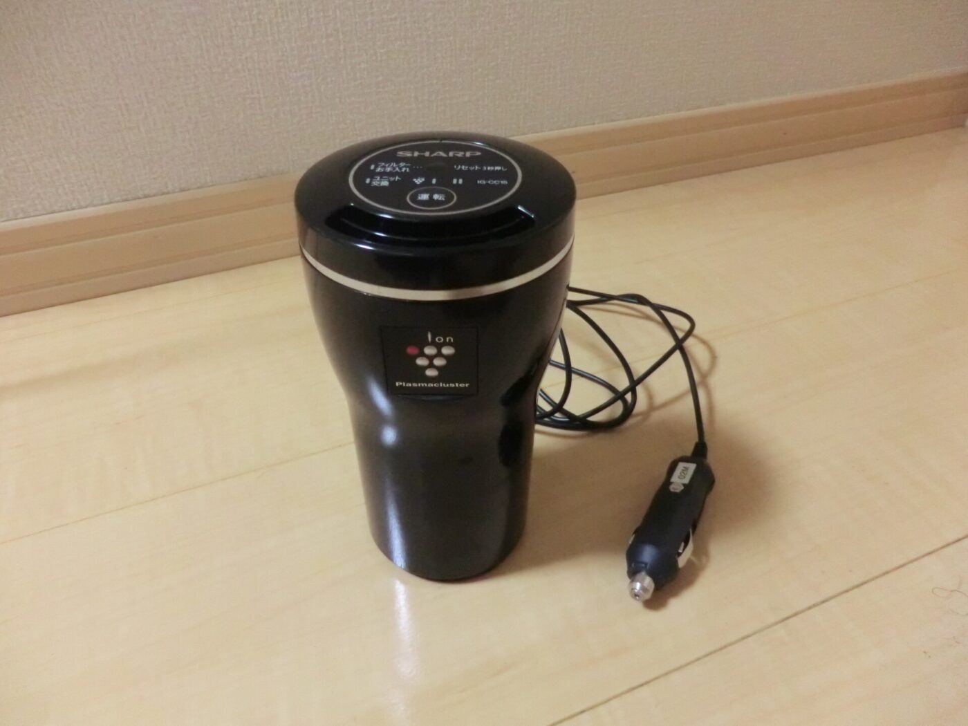 A cupholder-shaped car air purifier on a wood floor, with its cord and car socket plug.