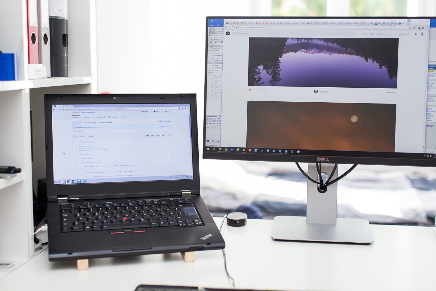 A laptop raised on a stand next to an external monitor on a white desk.
