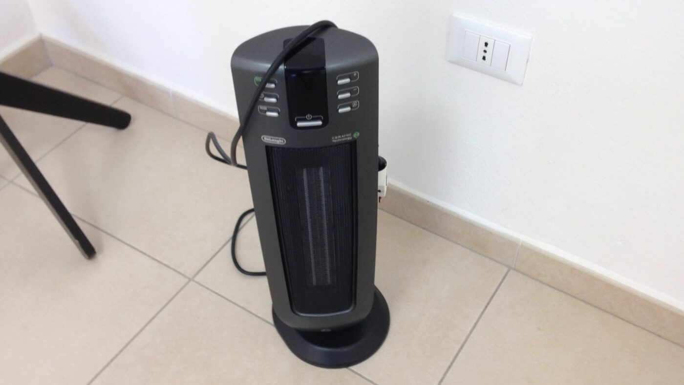 A slim tower heater standing on a tiled floor beside a wall outlet.