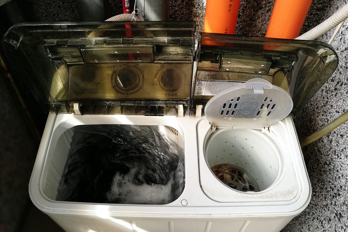 A twin-tub portable washer running a dark load, with hoses running to the wall behind it.