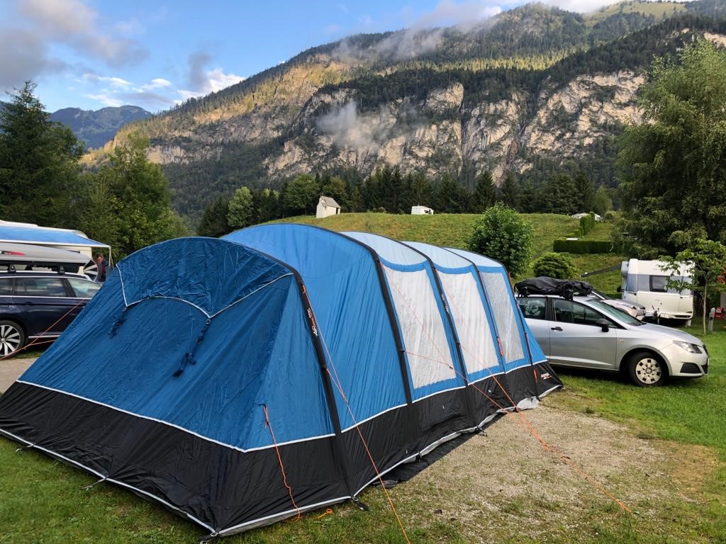 A large family tent pitched on grass at a mountain campground, with parked cars behind it.