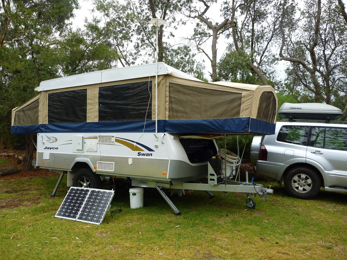 A small folding solar panel propped on the grass beside a pop-up camper trailer.