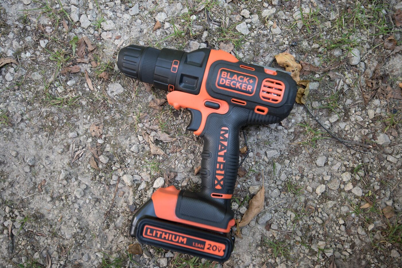 Orange and black cordless drill with a 20V lithium-ion battery pack lying on gravel