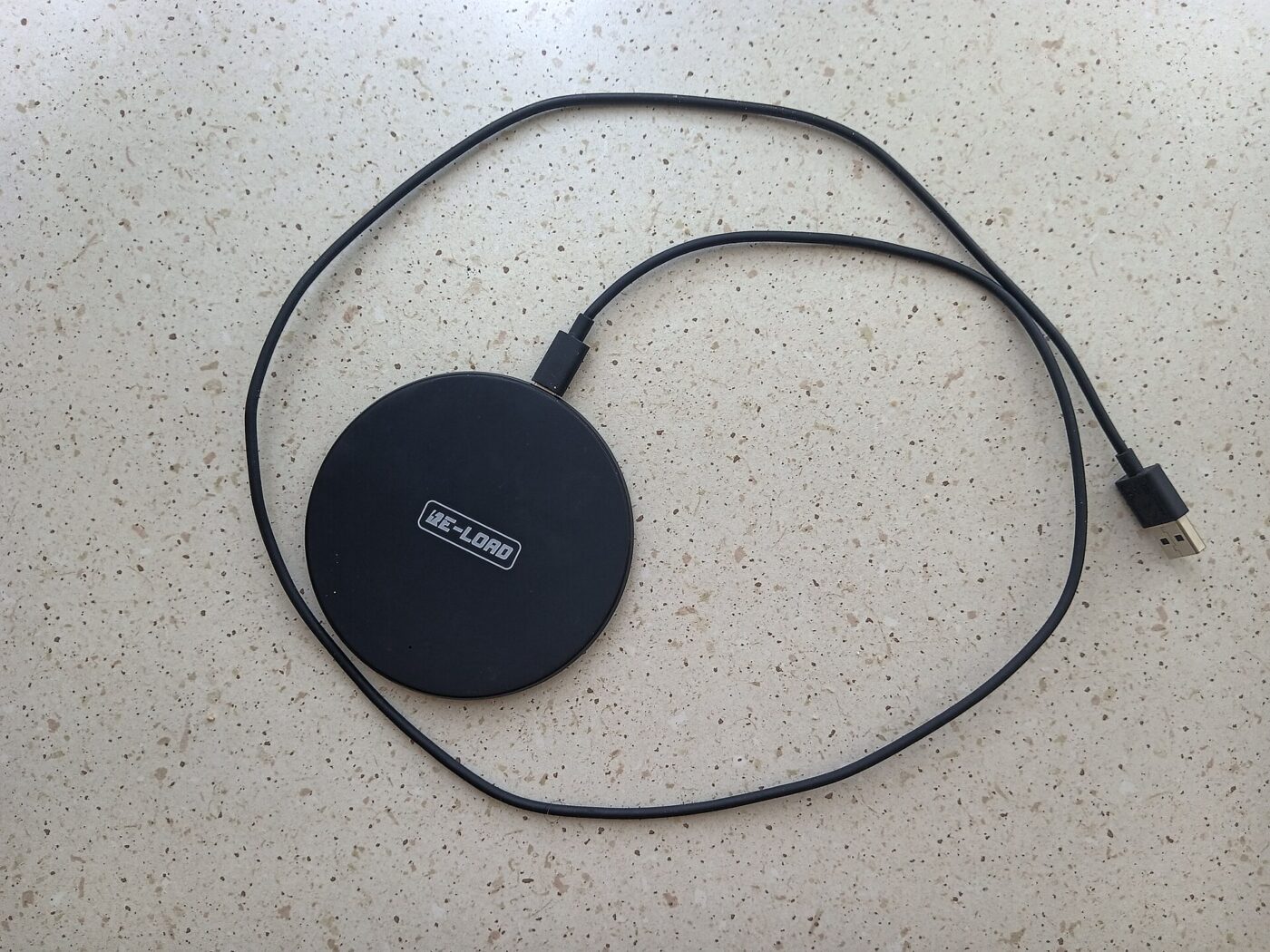 Flat black wireless charging pad with its USB cable coiled on a speckled countertop