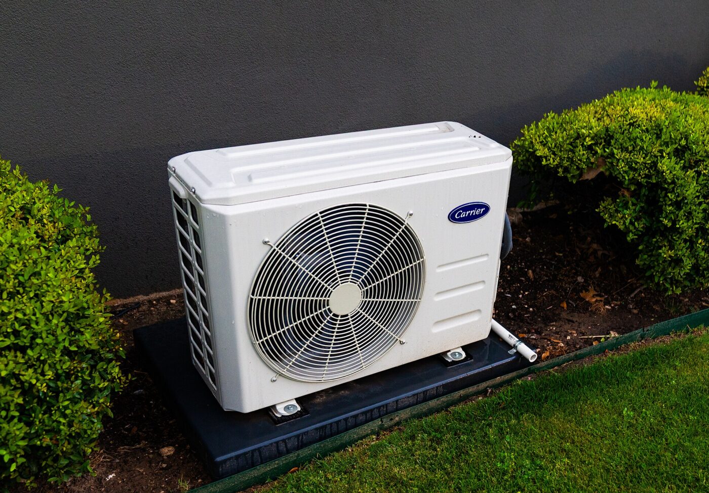 White mini-split heat pump outdoor unit on a dark pad between trimmed shrubs beside a lawn
