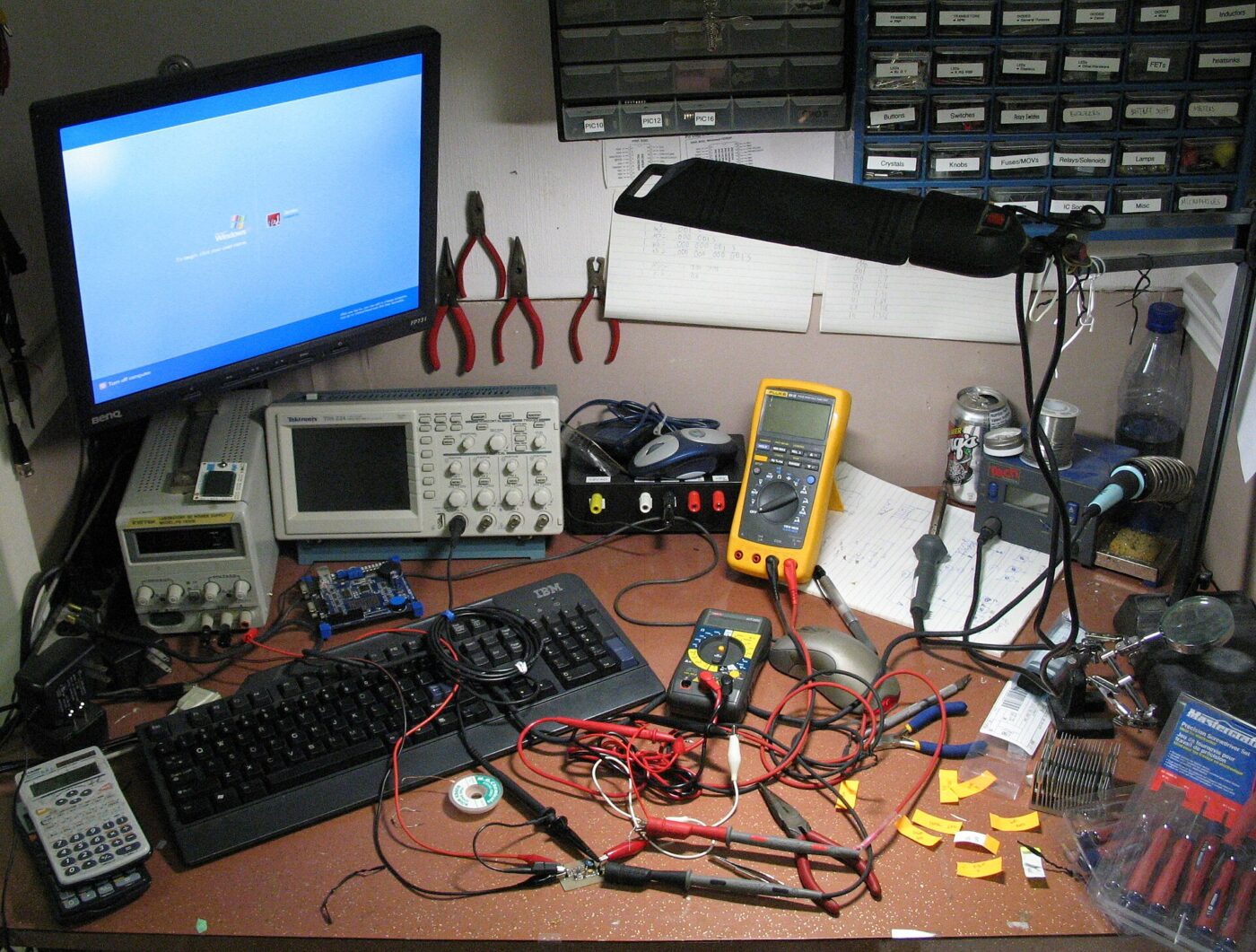 An electronics workbench with a multimeter, oscilloscope, soldering station, pliers and a tangle of test leads