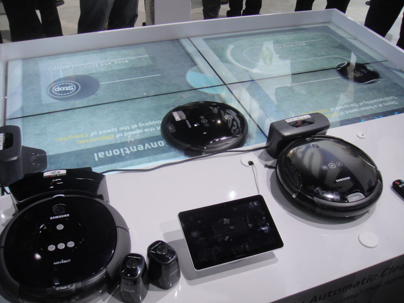 Three black robot vacuums, charging docks and a tablet on a trade-show demonstration table