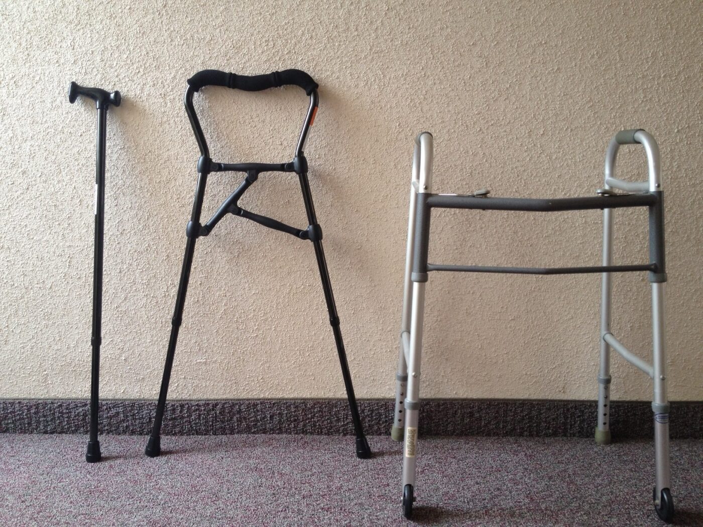 A walking cane, a four-legged cane hybrid and a folding walker frame stand against a wall.