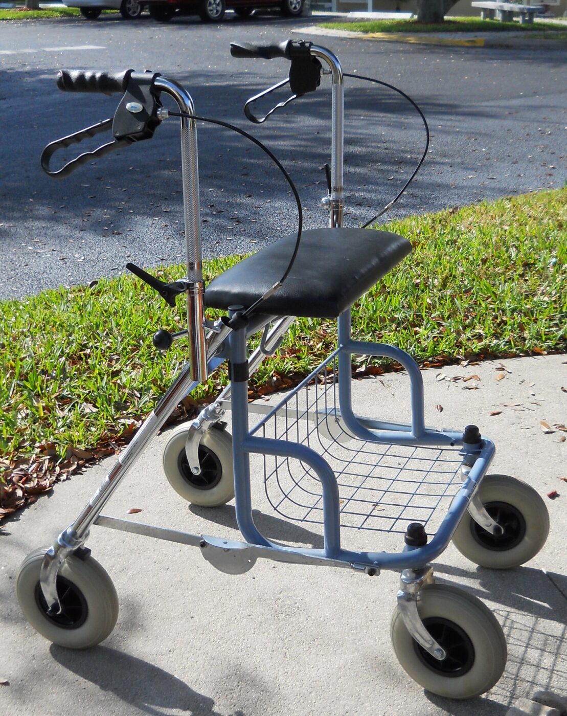 A four-wheel rollator with hand brakes, a padded seat and a wire basket on a sidewalk.