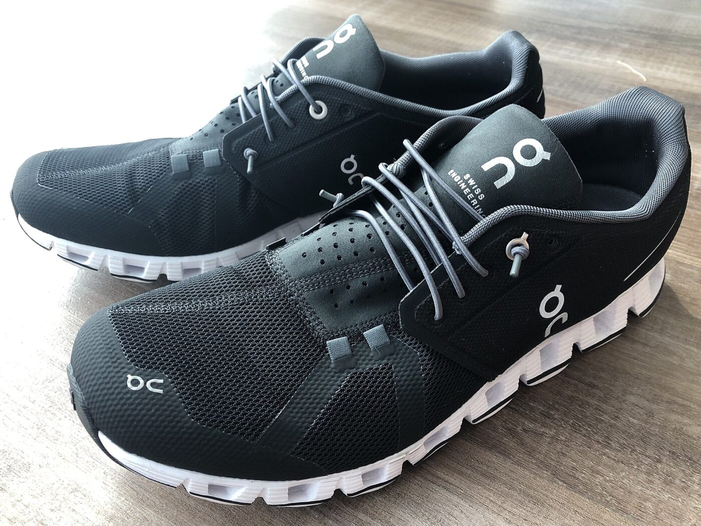 A pair of black On running shoes with white soles and gray laces on a wooden floor