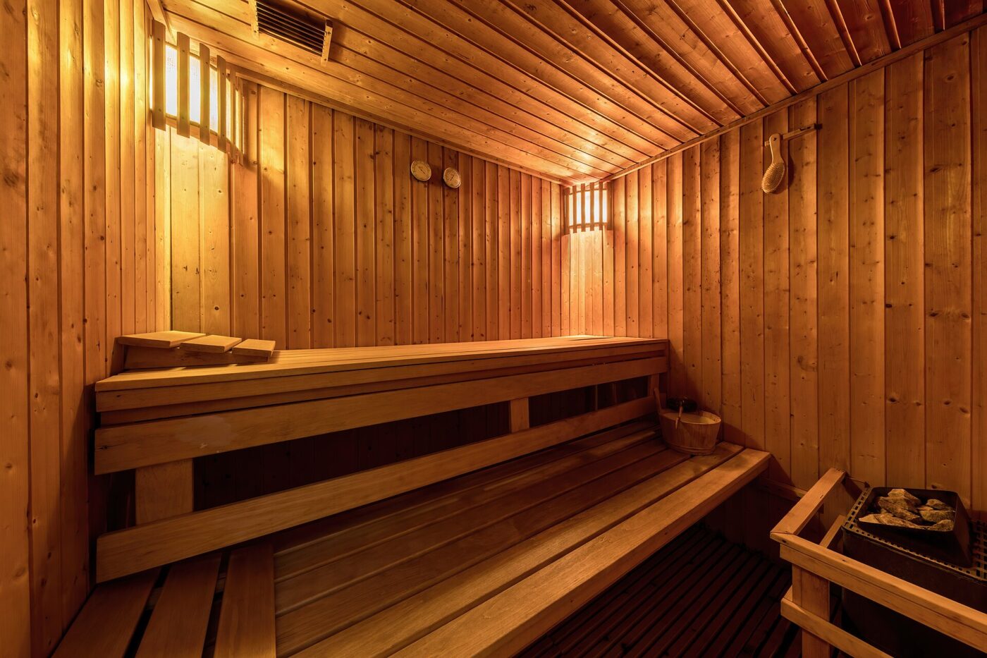A wood-paneled sauna with two tiers of benches, wall lamps and a stove with stones in the corner