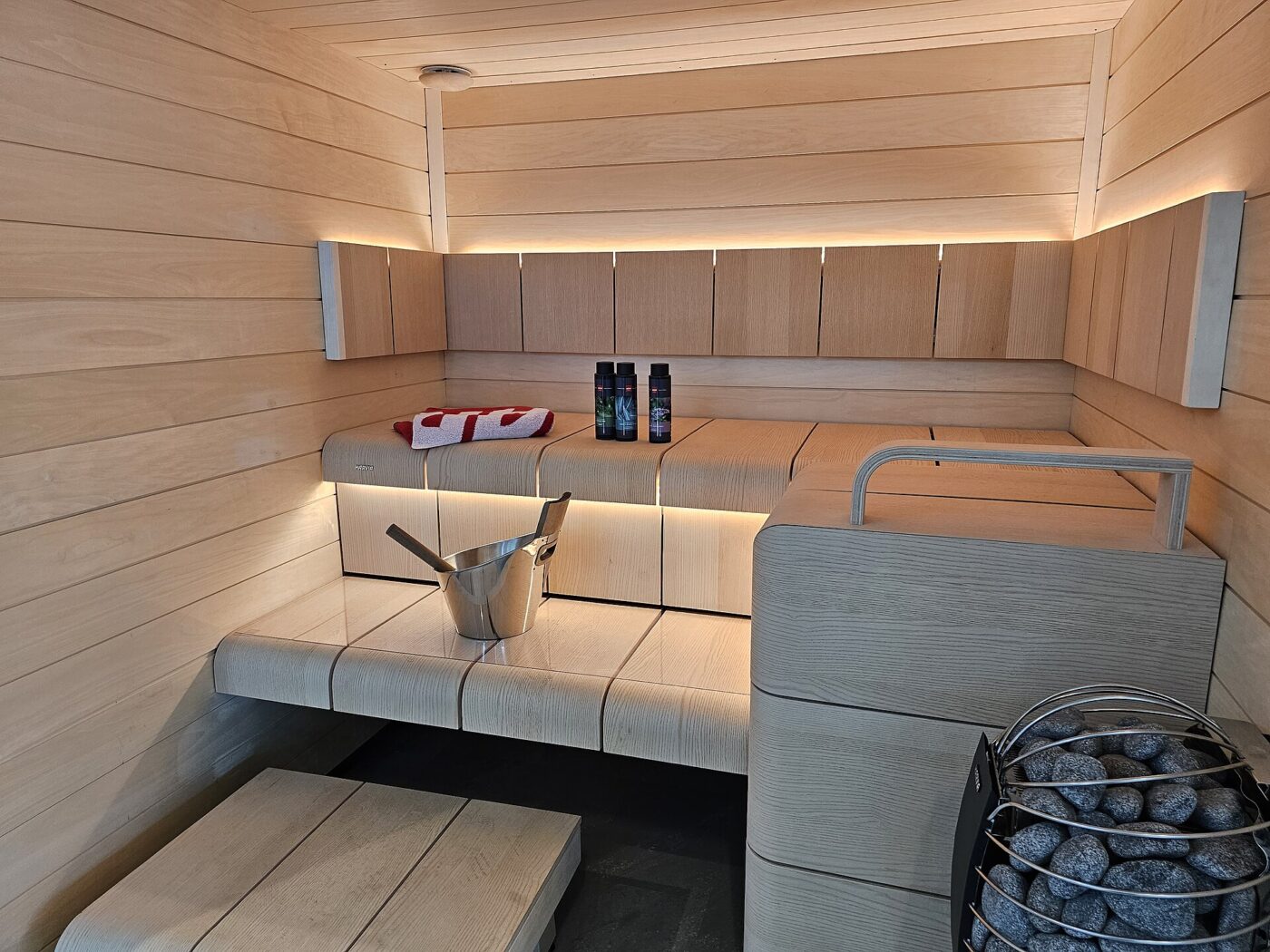 A modern sauna room with pale wood benches, strip lighting and a stone-filled heater in the corner
