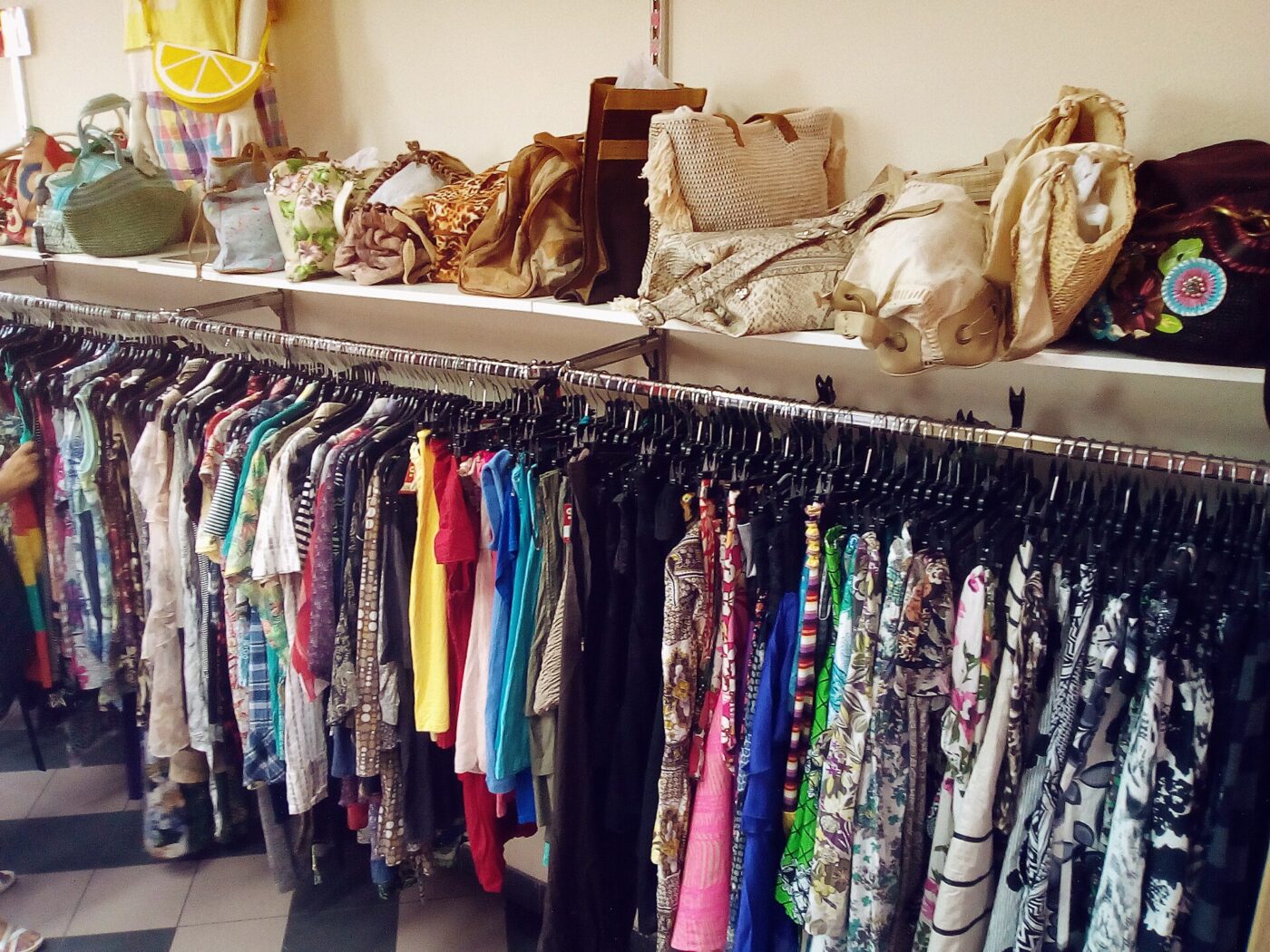 Racks of secondhand clothing under a shelf of used handbags in a thrift shop