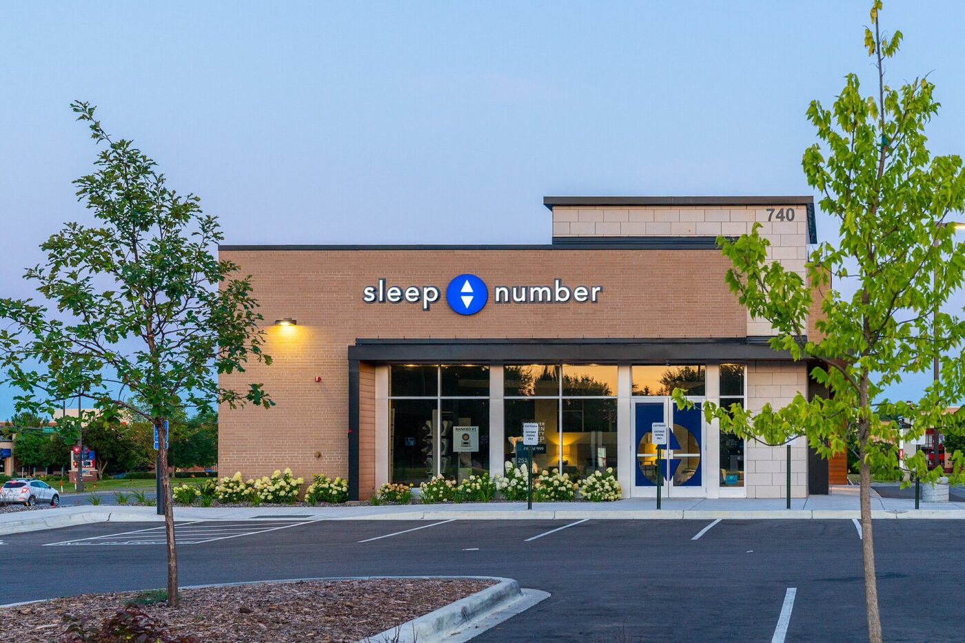 A Sleep Number store with lights on inside, facing an empty parking lot in the evening