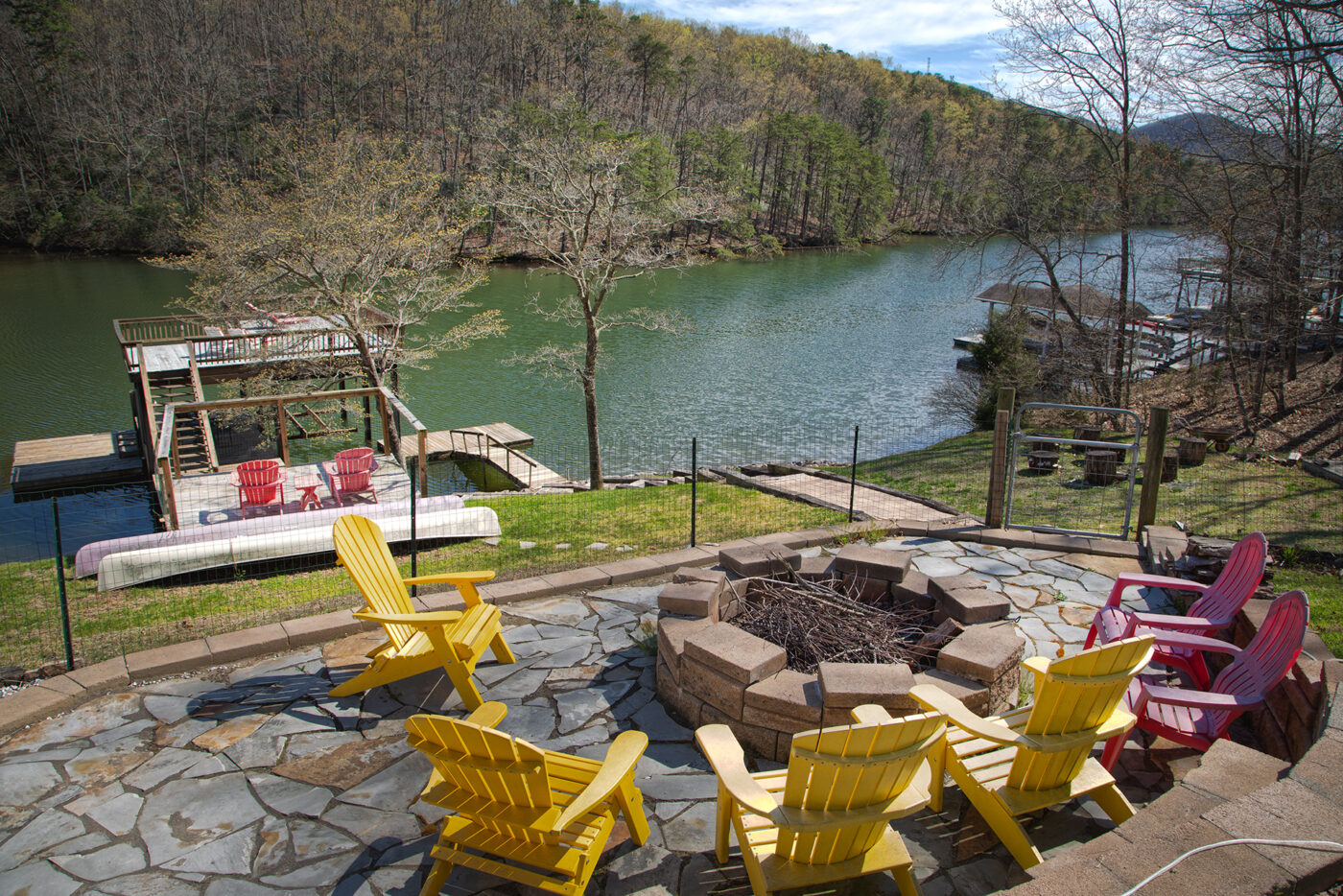 Adirondack chairs arranged around a stone fire pit on a lakeside backyard patio