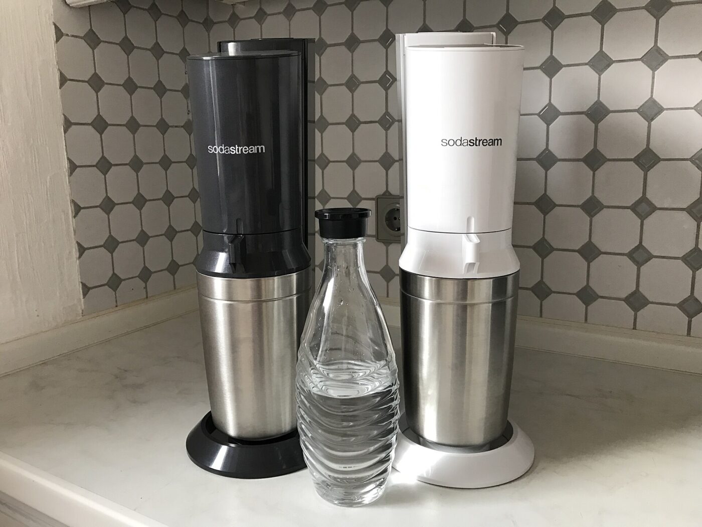Two countertop sparkling water makers standing beside a glass carafe of water on a kitchen counter.