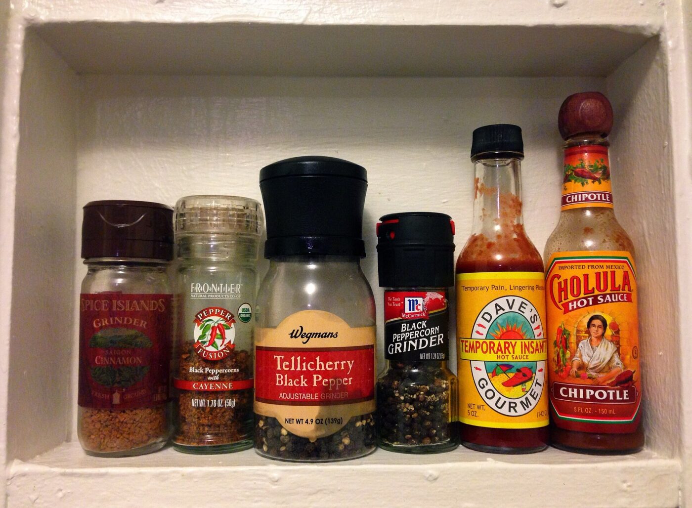 Store-bought spice jars and pepper grinders on a shelf beside two bottles of hot sauce