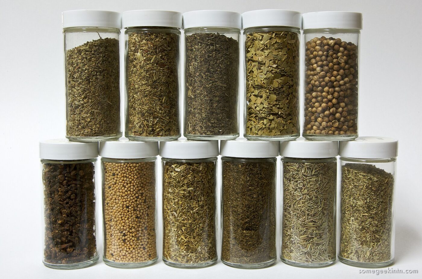 Eleven identical glass jars holding different whole and ground spices, arranged in two rows