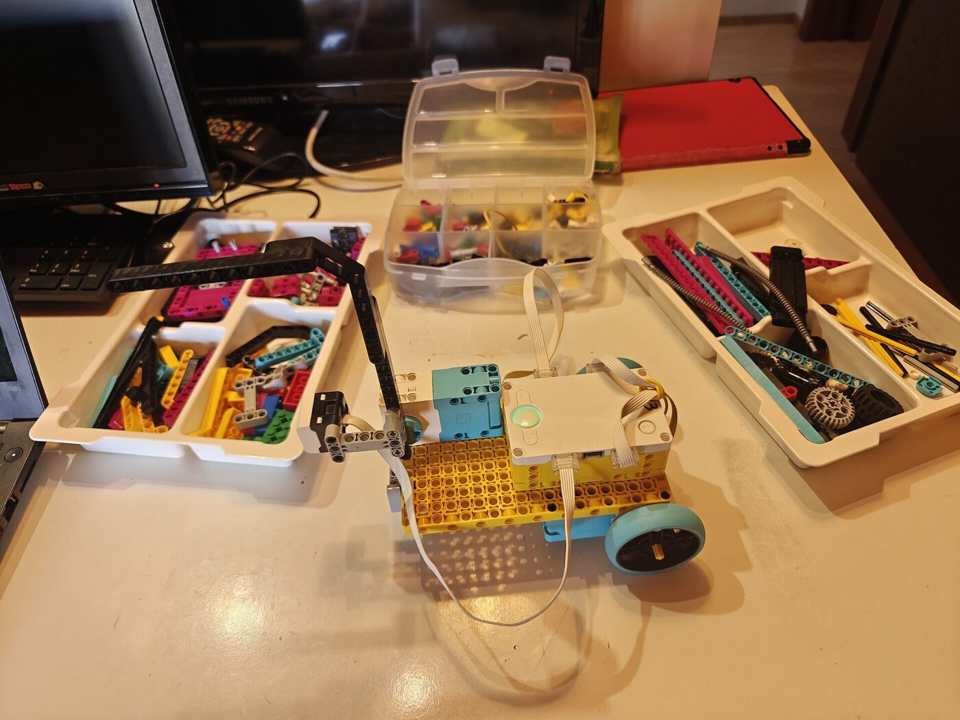A small brick-built robot on a desk beside trays of sorted beams, gears and wheels