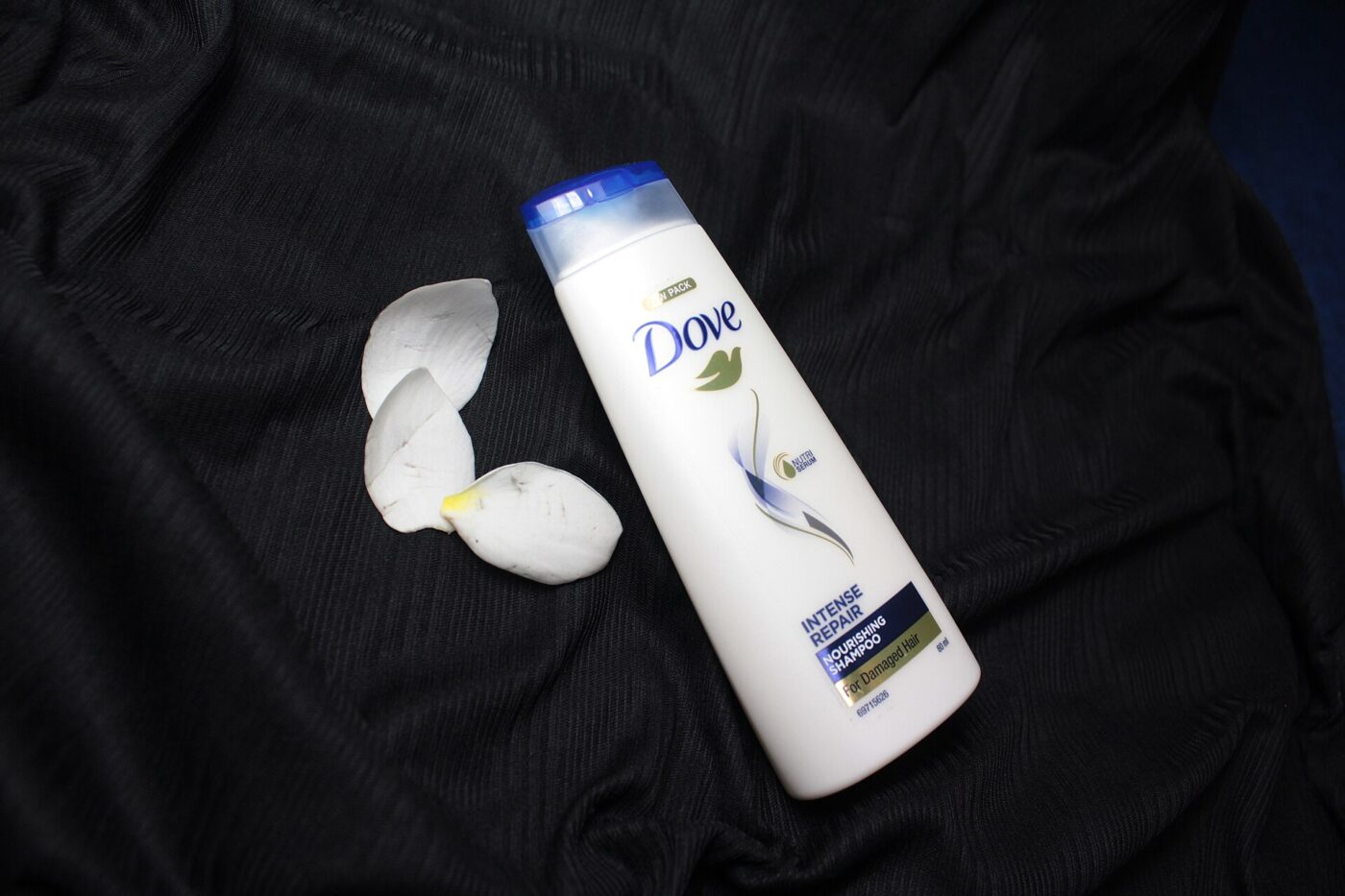 A white shampoo bottle lying on black fabric beside a few white flower petals