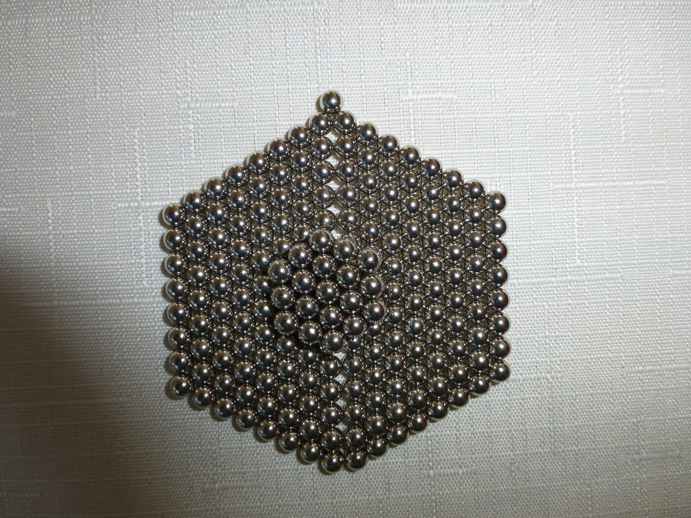 Hundreds of small steel magnet balls stacked into a cube on white fabric.