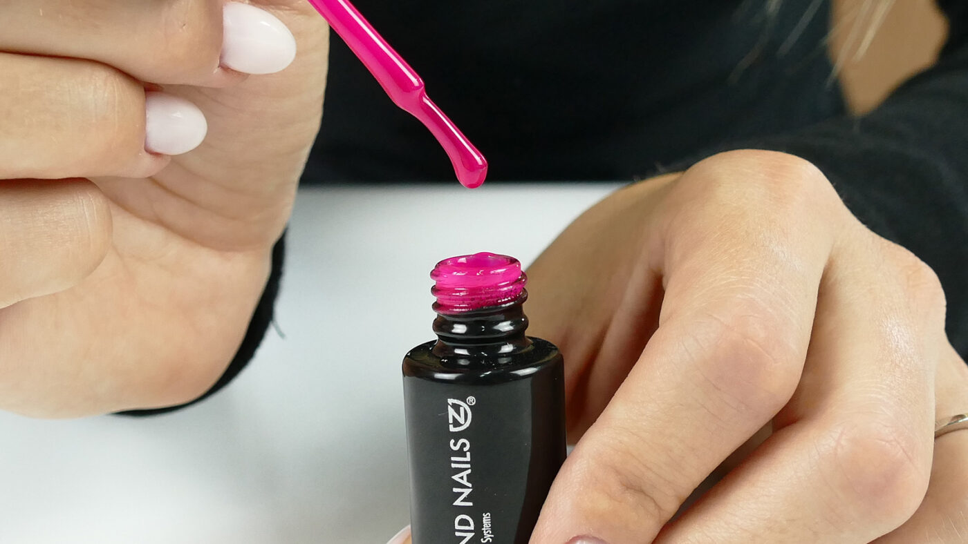 Hands hold an opened gel polish bottle and a brush above a white surface.