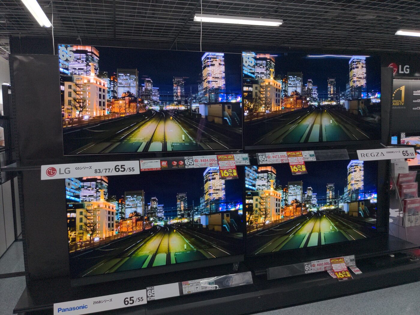 Four large televisions on store shelves showing the same night city scene above price cards.