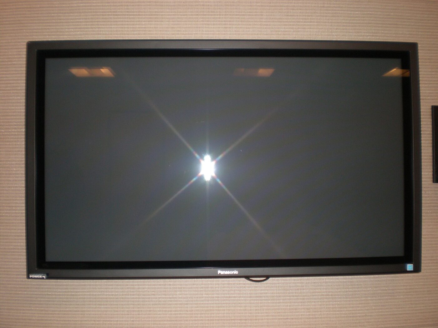 A flat-panel television mounted on a beige wall, screen off and reflecting a ceiling light.