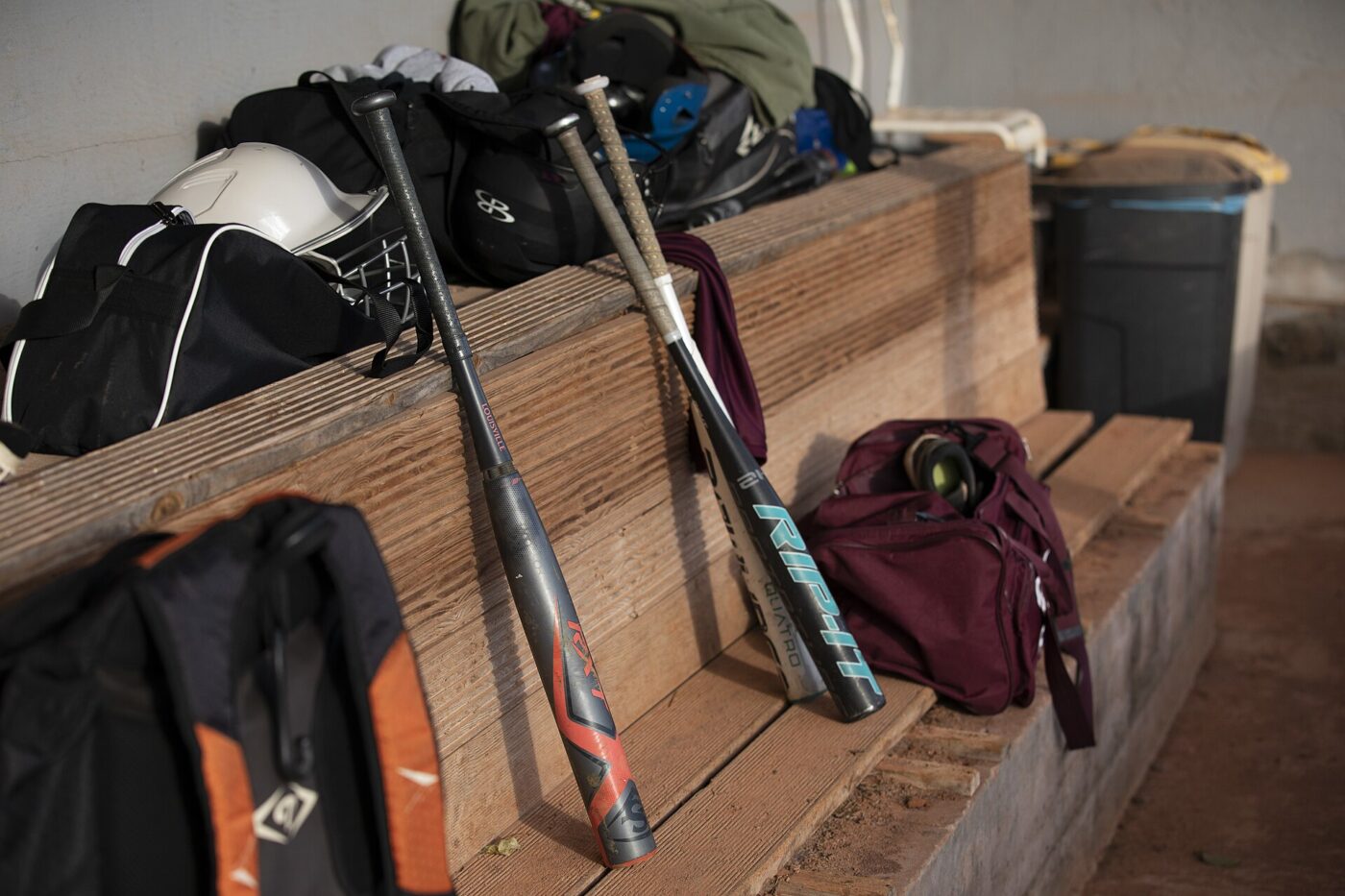 Two metal baseball bats lean against a wooden dugout bench next to gear bags.