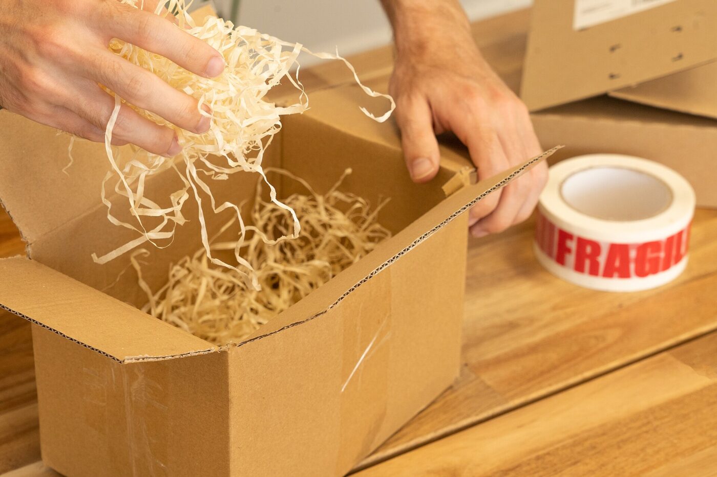 Hands fill a cardboard shipping box with wood wool packing beside a roll of fragile tape