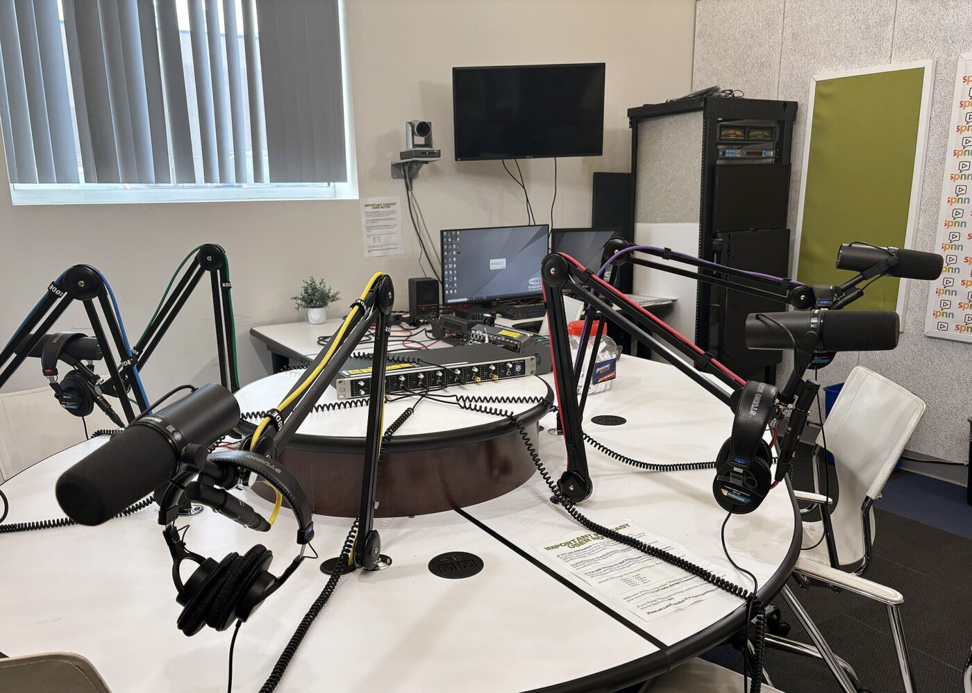 A podcast studio with four microphones on boom arms, headphones and a small camera mounted below a wall screen
