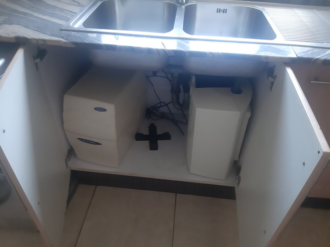 An under-sink water filter system installed in an open kitchen cabinet below a stainless steel double sink