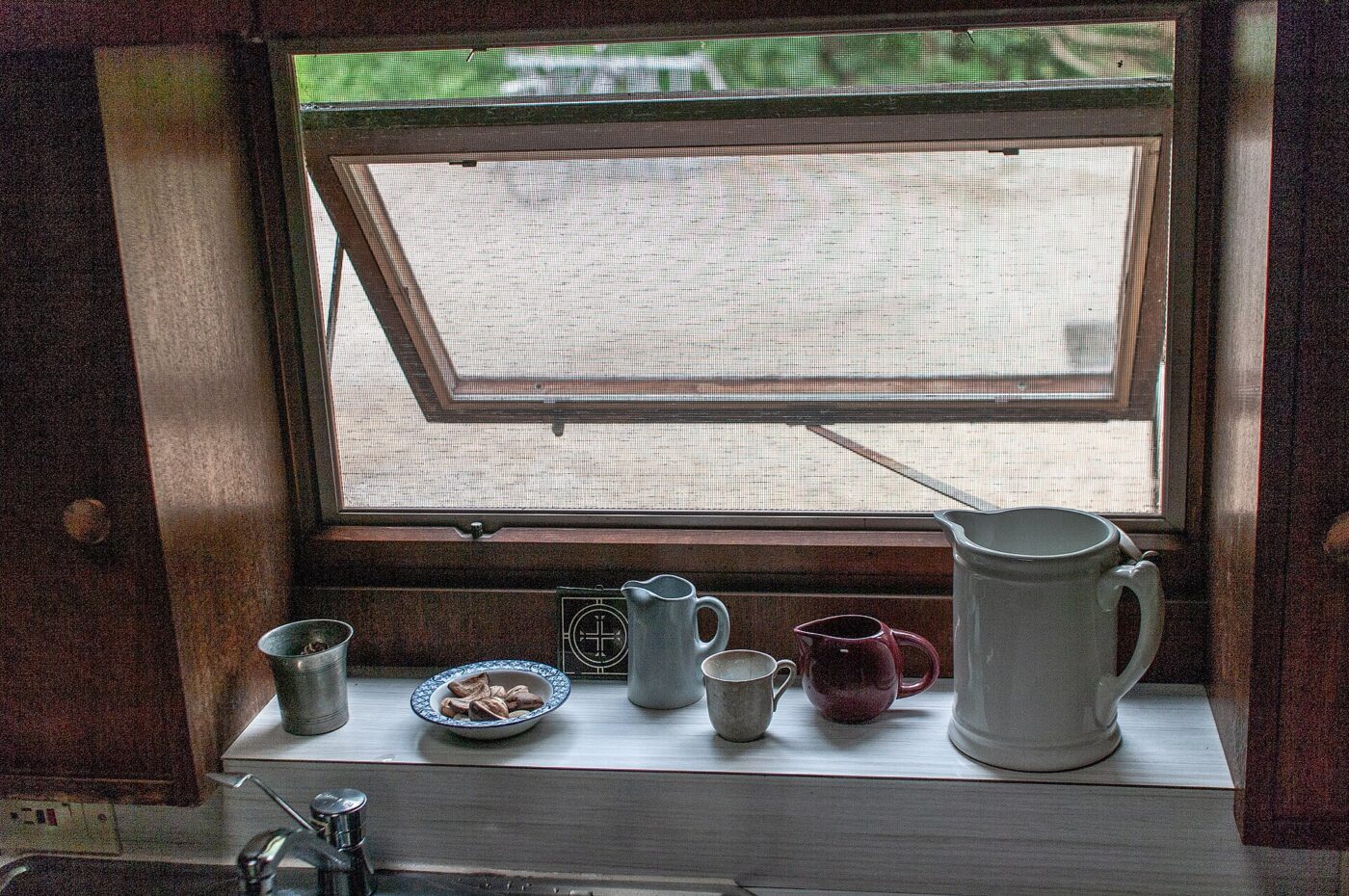 A kitchen window propped open above a sill holding pitchers, cups and a small plate.