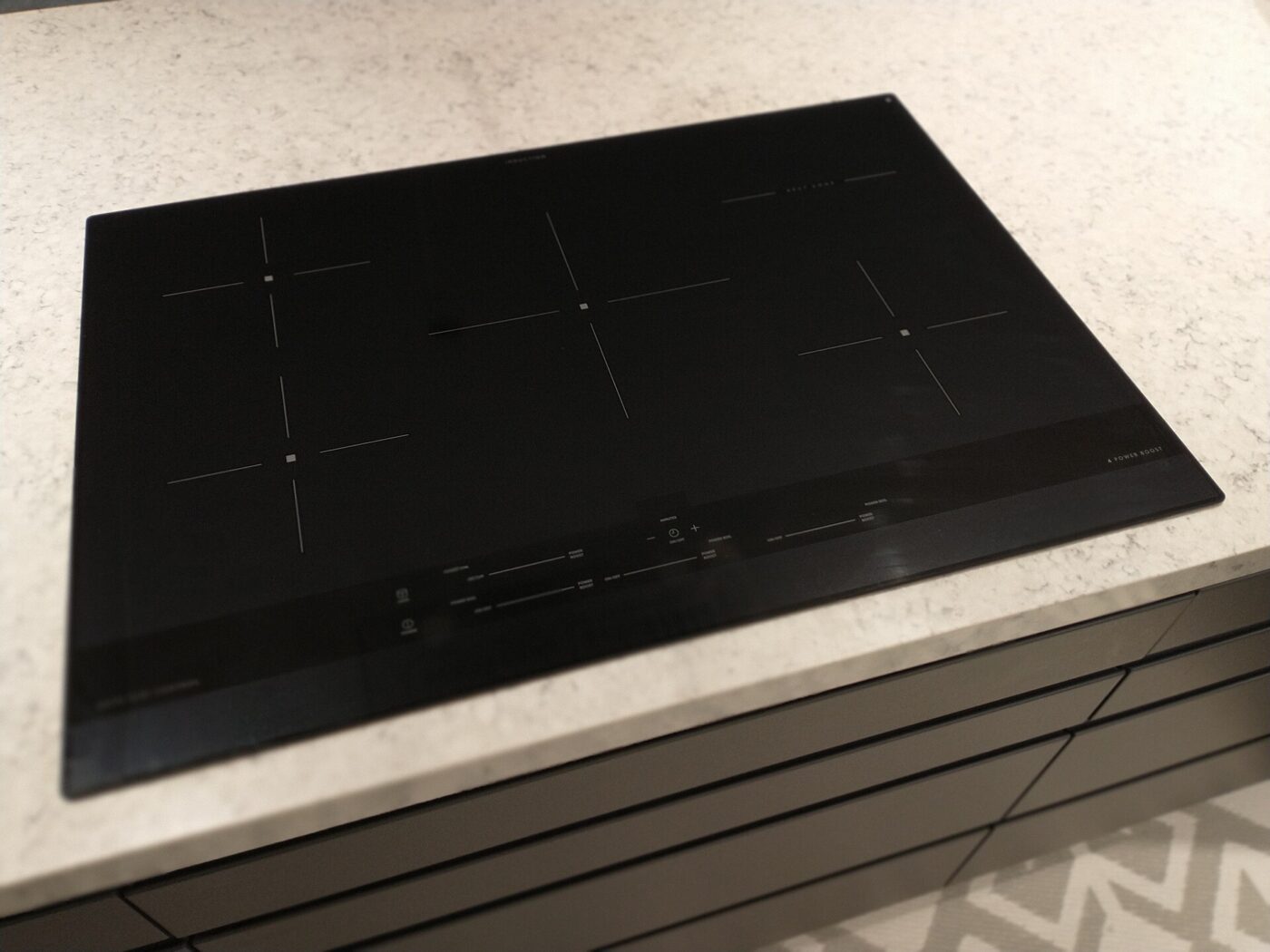A four-zone induction cooktop set into a pale stone countertop above dark cabinet fronts