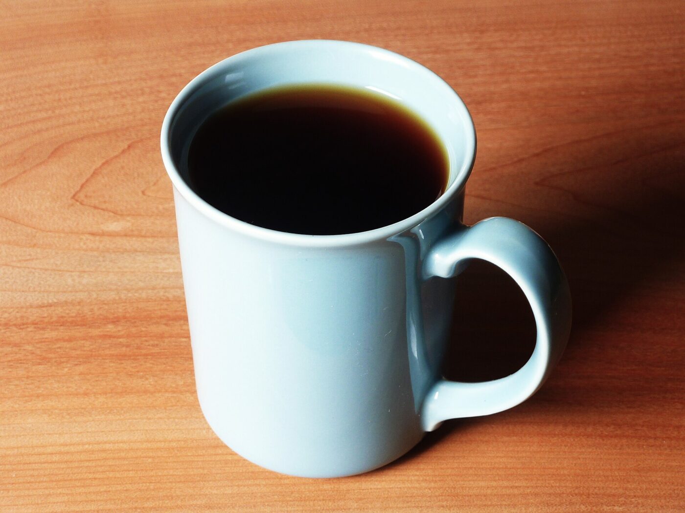 A pale blue ceramic mug of black coffee standing on a wooden desktop