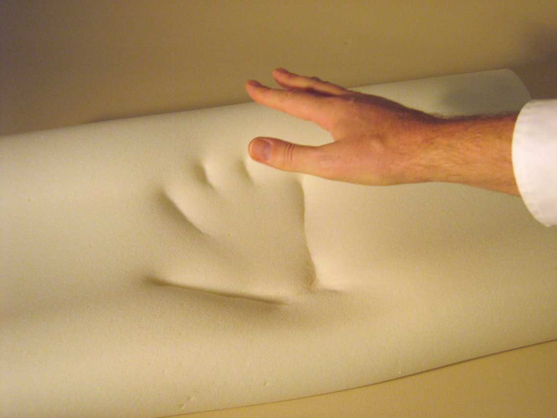 A hand pressing into a thick slab of memory foam leaves a deep, slowly recovering impression.
