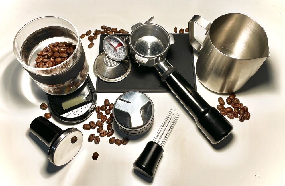 A portafilter, tamper, milk pitcher, scale and scattered roasted coffee beans on a pale surface.