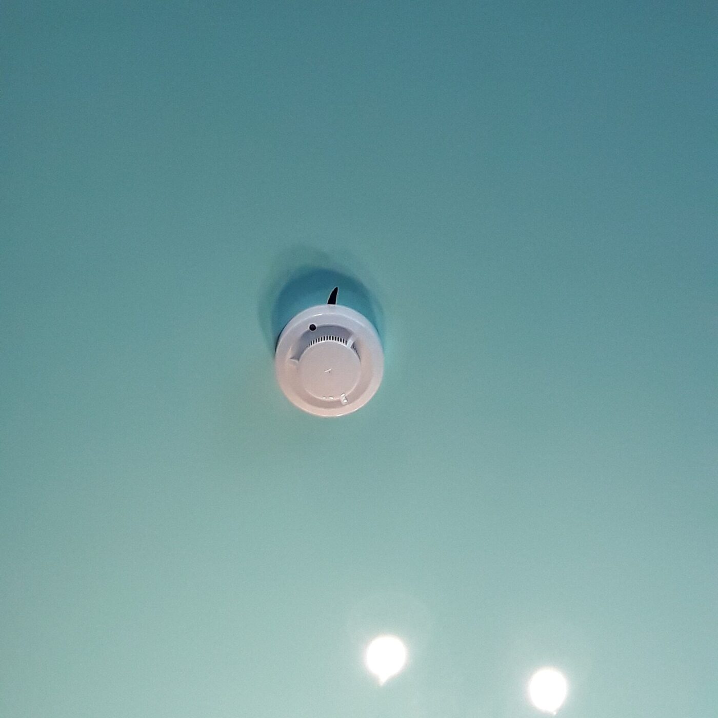 A white smoke alarm on a pale blue-green ceiling, with two recessed lights below