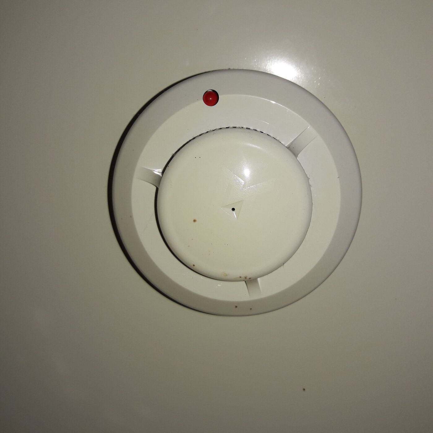 A round white smoke alarm fixed to a ceiling, its small red indicator showing