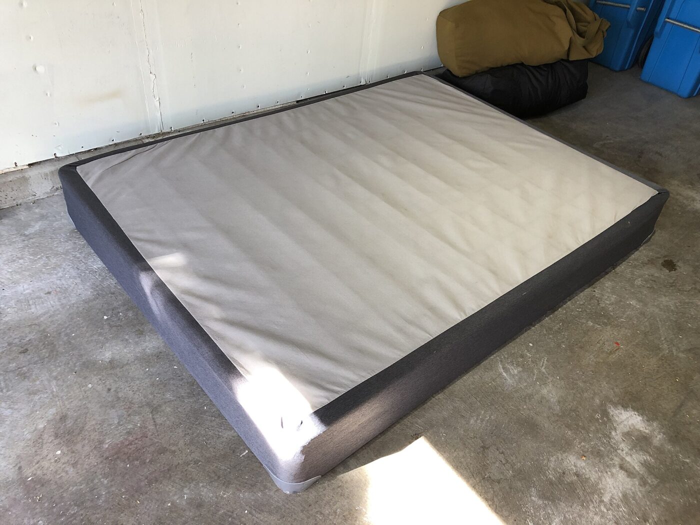 A fabric covered mattress foundation standing alone on a concrete floor, with no mattress on it.